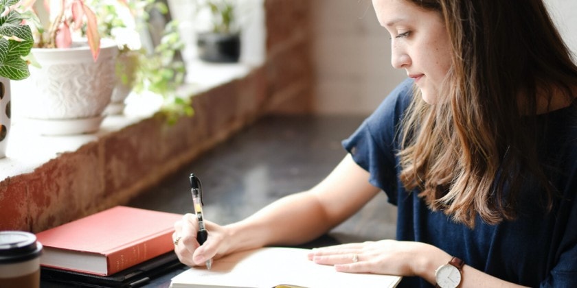 brunette girl writing in a book on a desk by a window
