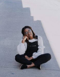 Girl with glasses and black overalls with legs crossed on the ground